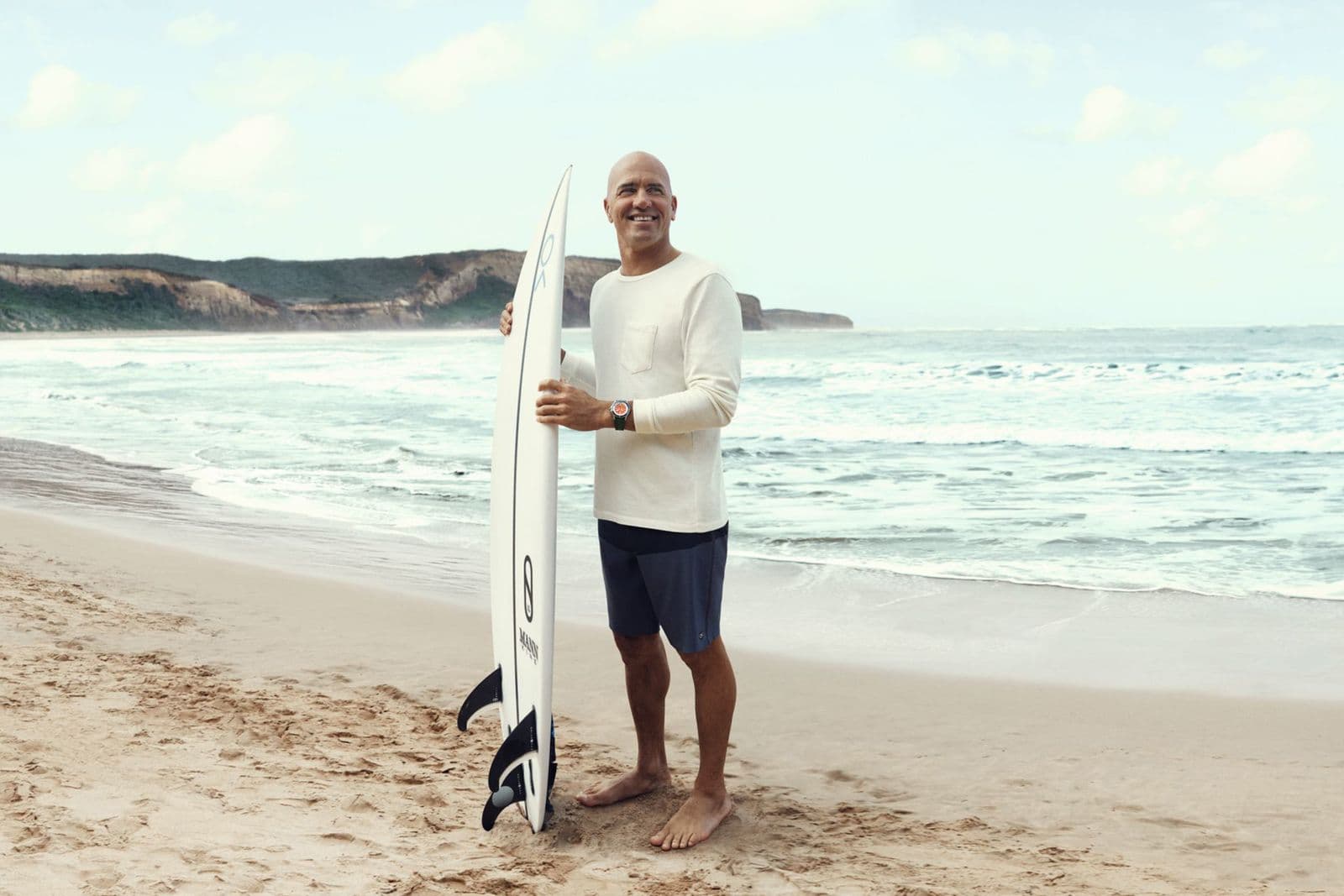 Kelly Slater holding a surfboard on the beach. Image credited to Breitling.